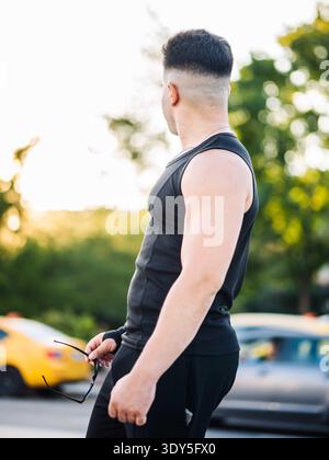 Jeune homme portant un débardeur et tenant des lunettes de soleil sur la rue urbaine au coucher du soleil Banque D'Images