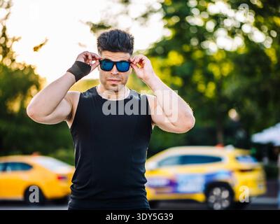Fit homme portant des lunettes de soleil et débardeur noir debout à l'extérieur dans le parc de la ville Banque D'Images