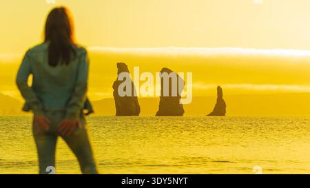 Une femme floue par derrière admire une vue sereine sur l'océan avec des piles de mer silhouettes. Tout le paysage est trempé dans une lumière jaune dorée, évoquer Banque D'Images