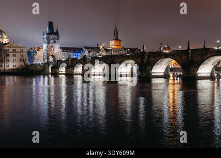 Vue nocturne hivernale du pont Charles à Prague photographiée depuis la rive de la petite ville. Les lumières douces de la ville se reflètent sur la rivière Vltava alors que les touristes traversent le pont historique Banque D'Images