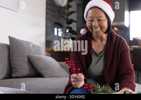 Femme asiatique senior portant un cardigan rouge, chapeau de Père Noël arrangeant la verdure sur le canapé, espace de copie. Vacances, festif, confortable, décoration, saisonnier, chaleur, intérieur || autorisation du modèle Banque D'Images