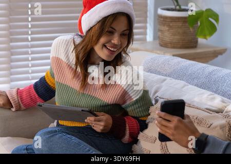 Couple assis sur le canapé à la maison, femme portant le chapeau de Père Noël tenant la tablette ; partenaire tenant le smartphone Banque D'Images
