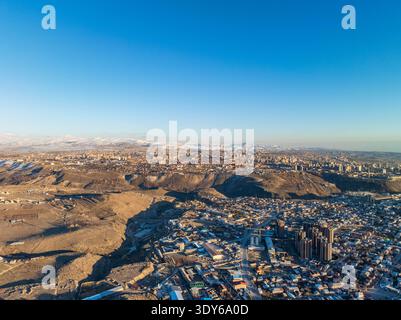Vue aérienne de la ville tentaculaire nichée entre collines ondulantes et montagnes lointaines. Ciel bleu clair au-dessus du paysage urbain avec résidentiel mixte et com Banque D'Images