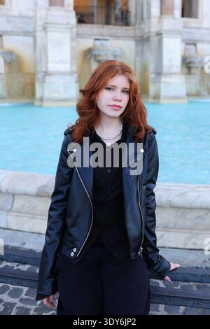 Belle femme rousse posant près d'une fontaine historique dans une place de la ville européenne. Banque D'Images
