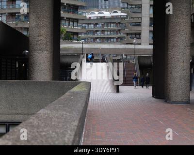 Les piétons montent la rampe de béton de White Lyon court, un carrefour clé dans le système de passerelles de haut niveau (Pedway) du Barbican Estate, Londres. En février 2026, cette artère reste un lien piétonnier essentiel reliant London Wall et Moorgate au Barbican Centre et à l'entrée de Silk Street de la Guildhall School of Music & Drama Banque D'Images