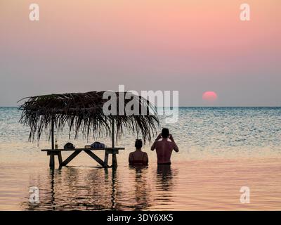 Vue sur le coucher du soleil au Swing Bar avec deux personnes se relaxant dans l'eau à secret Beach dans le district de Belize, Belize Banque D'Images