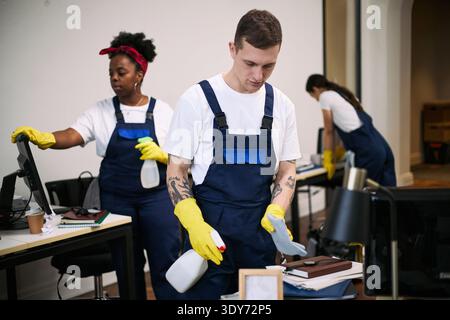 Jeune homme caucasien adulte nettoyant le bureau avec le flacon pulvérisateur et le chiffon tandis que jeune femme noire adulte et jeune femme adulte, travaillant en arrière-plan portant des uniformes de nettoyage et des gants Banque D'Images