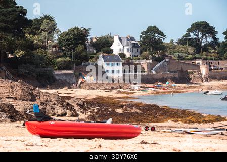 Le canoë rouge vif repose sur une plage rocheuse avec des maisons côtières et des pins à Rotheneuf Bretagne France Banque D'Images