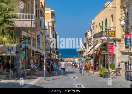 Vue sur la rue ensoleillée menant au port d'Héraklion, Crète avec des magasins et des gens appréciant la journée Banque D'Images