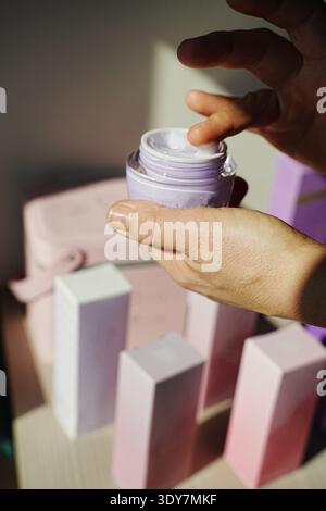 Gros plan des mains prélevant de la crème pour le visage dans un pot ouvert. L'image présente des produits de soins de la peau et des emballages, suggérant une routine de soins personnels axée sur Banque D'Images