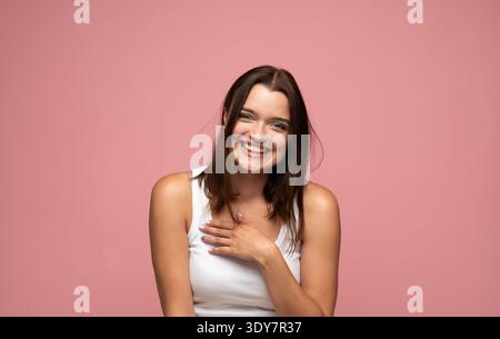 Jeune femme brune riant et exprimant la joie sur un fond rose, se sentant positive et heureuse Banque D'Images