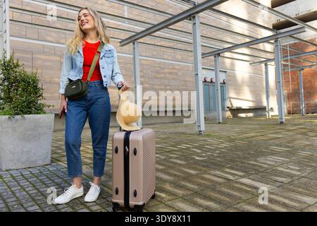Femme souriant dans la veste en denim à côté de la valise avec chapeau de paille, tenant le téléphone dans la plaza, espace de copie Banque D'Images