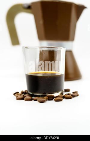 Verre avec espresso foncé sur la surface blanche, pot de moka flou en arrière-plan, grains de café dispersés autour Banque D'Images