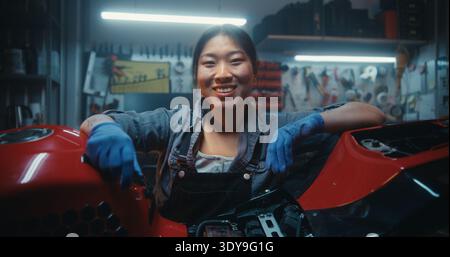 Portrait de mécanicien féminin heureux souriant se penchant sur la moto après réparation réussie. Satisfaction de jeune femme asiatique après avoir terminé l'entretien technique et la réparation électronique dans l'atelier de garage. Banque D'Images