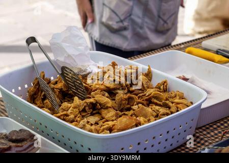 Un lot de croûtes de porc frites croustillantes dorées ou de chicharrones servies dans un panier en plastique blanc avec des pinces en métal sur un marché. Banque D'Images