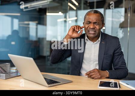 Homme d'affaires mature éprouvant de graves douleurs à l'oreille et un inconfort, touchant son oreille tout en travaillant sur un ordinateur portable à son bureau, indiquant des problèmes auditifs ou des acouphènes pendant une journée de travail chargée Banque D'Images