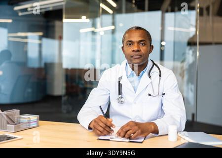 Médecin masculin professionnel en blouse de laboratoire et stéthoscope est assis à un bureau dans un bureau moderne, écrivant sur un presse-papiers et regardant avec confiance la caméra, symbolisant la confiance et l'expertise médicale Banque D'Images