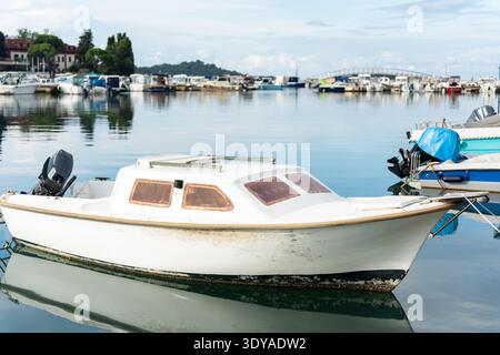 Petit bateau à moteur blanc vieilli avec moteur hors-bord flottant dans les eaux calmes du port avec une marina bondée. Banque D'Images
