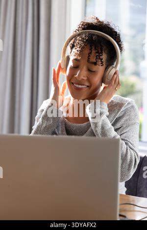 Femme afro-américaine assise à table portant un pull gris clair, tenant des écouteurs et un ordinateur portable Banque D'Images