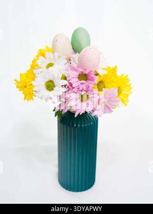 Un bouquet printanier festif de marguerites jaunes, blanches et roses avec des œufs de Pâques mouchetés est disposé dans un vase côtelé vert foncé sur une table blanche. Banque D'Images