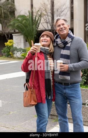 Couple senior debout sur la place portant un bonnet et des foulards tenant des tasses à café pointant vers le haut Banque D'Images