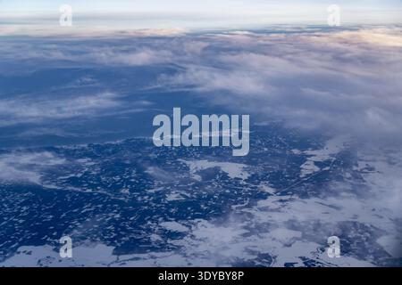 Panorama aérien de terres enneigées et de forêts vu à travers des couches de nuages brisées, des tons bleus froids et une brume douce à l'altitude de croisière, persp. De vol atmosphérique Banque D'Images