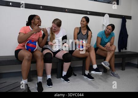 Diverses coéquipières féminines de volley-ball assis sur un banc en bois dans le vestiaire contenant des balles de volley-ball. Équipe, balle, siège, camaraderie, athlétique, occasionnel, multiculturel || autorisation du modèle Banque D'Images