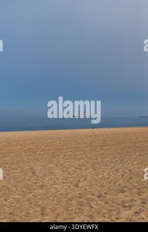 Large plage de sable rencontre une mer bleu ardoise calme sous un ciel brumeux. Un navire industriel avec des grues jaunes ancres offshore, avec des marqueurs de canal et un dist Banque D'Images