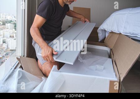 Homme assemblant et déballant de nouveaux meubles d'emballage plat d'une boîte en carton dans une chambre à coucher Banque D'Images