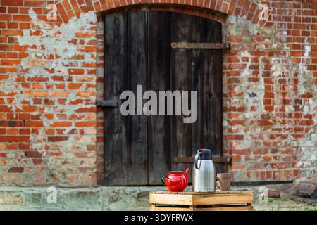 Une scène extérieure rustique avec une théière rouge, un thermos en acier inoxydable et une tasse en céramique placée sur une palette en bois devant une ancienne porte de ferme. Banque D'Images