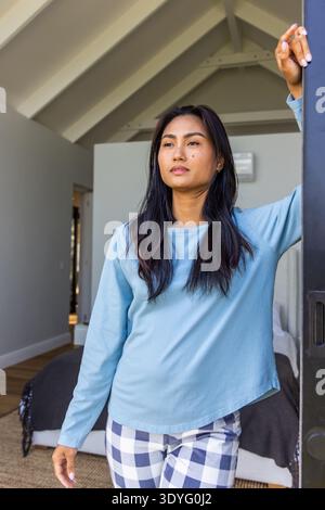 Femme asiatique portant un haut bleu clair, un pantalon à carreaux, appuyée sur un cadre de porte sombre dans la chambre, montrant le lit Banque D'Images