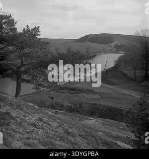 Photographie vintage en noir et blanc offrant une vue panoramique panoramique surélevée d'une vaste vallée fluviale pendant la saison d'automne. Un chemin de terre longe la côte herbeuse accidentée où une moto soviétique classique est stationnée tranquillement, surplombant de petits bateaux en bois amarrés à proximité. Un majestueux pin encadre la gauche, tandis qu'un pont étroit est visible au loin. Ce paysage très détaillé capture magnifiquement les loisirs ruraux quotidiens du milieu du XXe siècle, servant de fenêtre nostalgique sur un Donbass paisible avant la guerre. Banque D'Images