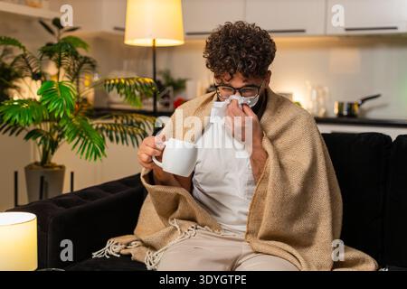 L'homme arabe à la maison se sent mal à la gorge tient tasse de thé chaud avec la médecine prend lente gorgée utilise le tissu. Égyptien sur le canapé fouille médecin sur smartphone frotte toux cou repose sous la fièvre sniff couverture Banque D'Images
