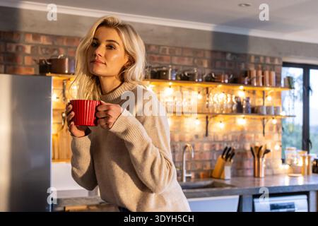 Femme à la fin des années 20 debout dans la cuisine à la maison portant un pull beige tenant une tasse côtelée rouge Banque D'Images