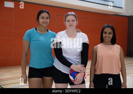 Diverses coéquipières féminines debout sur le terrain de gymnastique tenant le volley rouge blanc bleu devant le mur orange. Athlète, travail d'équipe, bois dur, lignes, rembourrage, gymnase, vibrant || autorisation du modèle Banque D'Images