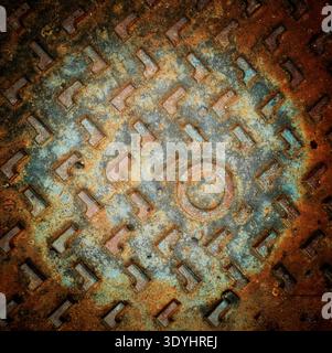 Le couvercle de trou d'homme en métal fortement corrodé détaille un riche mélange de rouille orange et d'oxydation vert bleu. Les motifs géométriques ajoutent de la texture à la vieille ville Banque D'Images