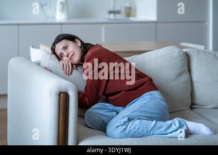 Femme calme se relaxant sur un canapé confortable à la maison, appuyée sur un oreiller avec un sourire paisible appréciant le confort. Banque D'Images