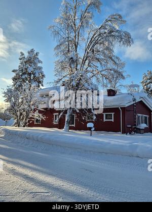 Explorez une scène sereine à Akaslompolo, en Finlande, où une charmante cabane rouge se dresse au milieu d'une couverture de neige. De grands arbres scintillent sous le SK bleu clair Banque D'Images