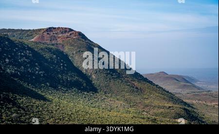 Large vue panoramique sur une crête montagneuse volcanique verte avec un bord de cratère rocheux sous la brume bleue du ciel, surplombant une vaste vallée et des plaines lointaines. Banque D'Images