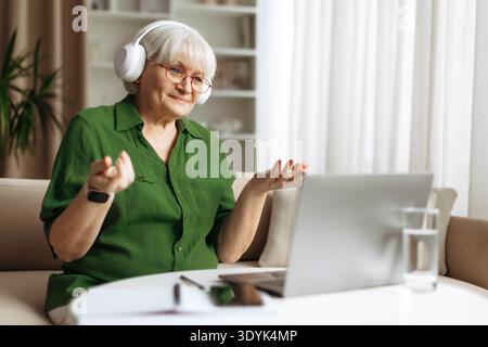 Femme senior portant des écouteurs et des gestes tout en faisant un appel vidéo sur ordinateur portable Banque D'Images