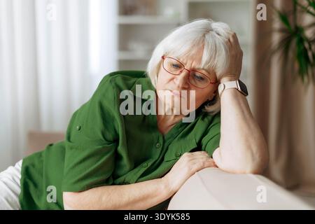 Femme senior assise sur le canapé tenant la tête avec une expression réfléchie à la maison Banque D'Images