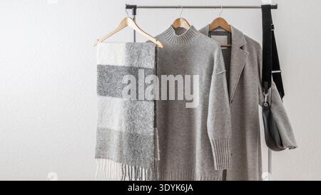 Manteau ou veste en laine gris clair, pull ou robe mérinos, écharpe en alpaga et sac en cuir noir sur cintre sur fond mural blanc. Automne et hiver Banque D'Images