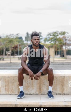 Corps complet de l'athlète masculin afro-américain en vêtements de sport assis à la frontière pendant l'entraînement de fitness et le repos Banque D'Images
