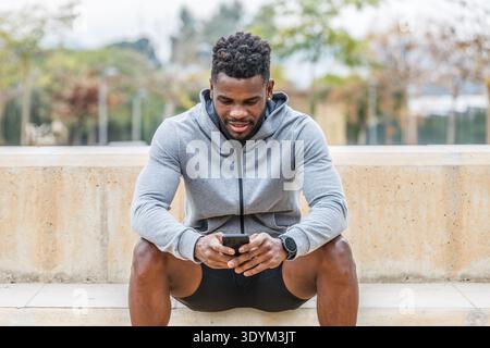 Jeune athlète masculin afro-américain dans des vêtements de sport assis sur les escaliers et utilisant le téléphone portable tout en surfant sur Internet pendant la pause de traini extérieur Banque D'Images