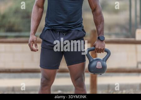 Recadrez l'athlète afro-américain anonyme avec un corps musclé sportif portant des vêtements de sport noirs serrés tenant une kettlebell lourde pendant un haltlifti intense Banque D'Images