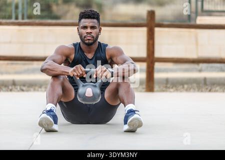 Corps complet d'athlète barbu confiant en vêtements de sport et en baskets assis et soulevant une kettlebell lourde pendant l'entraînement d'haltérophilie en plein air l'été Banque D'Images