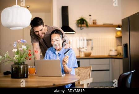 Un couple joyeux profite d'un moment agréable dans leur cuisine confortable, célébrant ensemble des nouvelles passionnantes tout en travaillant sur leur ordinateur portable. Banque D'Images