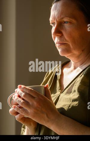 Femme d'âge moyen réfléchie tenant une tasse de thé par fenêtre dans la lumière chaude du matin, moment calme de réflexion à la maison. mise au point sélective Banque D'Images
