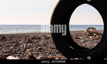 Déchets dispersés et objets cassés sur la rive rocheuse contre la mer bleue, vu à travers de gros pneus jetés. Banque D'Images