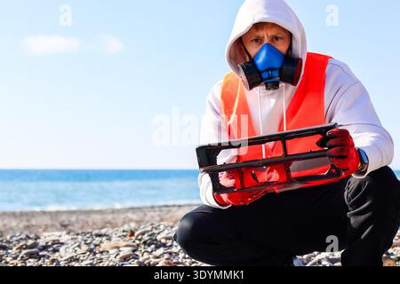 Figure squatting dans respirateur et gilet présente un récipient en plastique noir sur la plage remplie de galets au bord de la mer. Banque D'Images